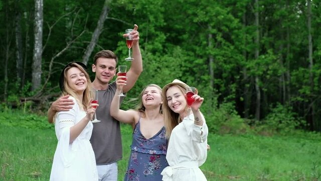 group cheerful laughing young friends enjoy picnic in forest in clearing on summer day. man and a woman dance outside the house with red wine drink a cocktail have fun spend active holidays in nature