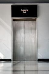 A stainless steel door of an elevator with refelcting tiles on the floor and a white wall.
