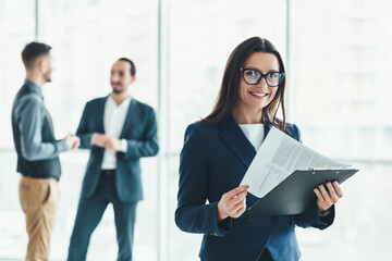 Young female assistand standing on the forefront with all the documents ready for signing, while two men are negotiating on the background.