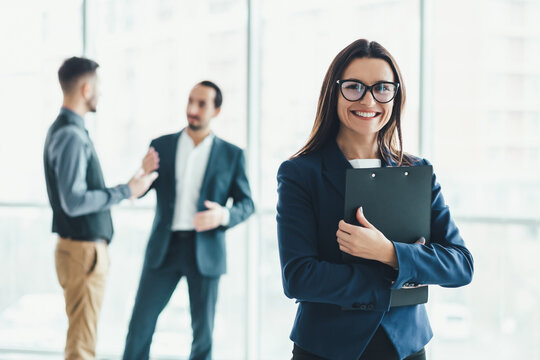 Portrait of pretty office female employee with folder in hands, smiling lovely at the camera and her coleagues discussing something on blurred background.