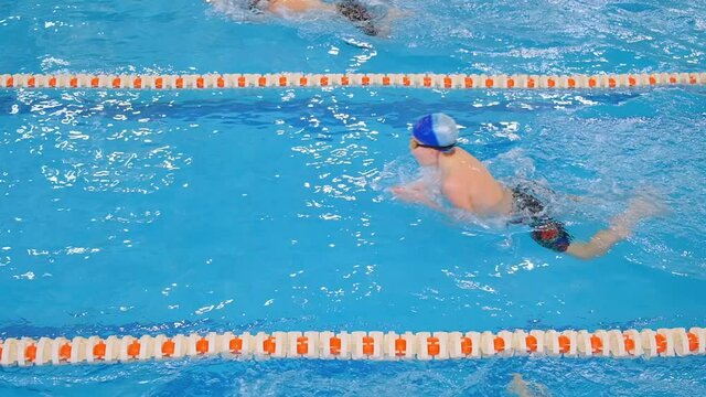 young competitors swim race in breaststroke in swimming pool between coloured ropes under bright electric light slow motion