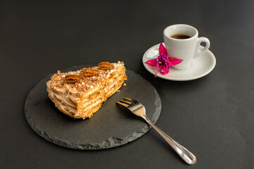 A typical Portuguese cake made of cookies "Maria" called Bolo de Bolasa with cream, pecans, black and white chocolate, on a black slate with a fork, with espresso and a flower on a black background