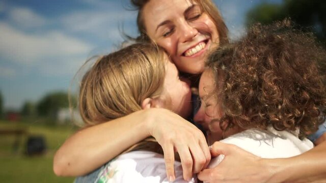 Cheerful mom and two daughters in white t-shirts hagging together outdoors. Mothers day, happy family on a sunny summer day