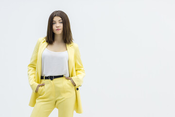 Cheerful beautiful girl holding hands in pockets of bright stylish yellow suit in studio on white background.