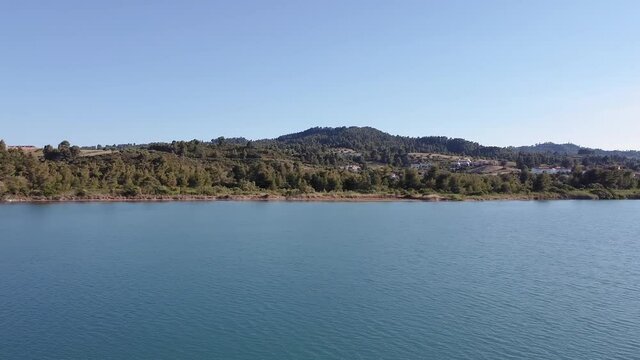 Drone flying at low altitude, with caique fishing boats at calm sea. Aerial top view of Glarokavos lagoon at Kassandra Chalkidiki peninsula shoreline with green plantation & pine-clad hill houses.