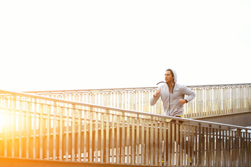 Sporty young man working out at early morning while run up the bridge with orange sunrise on background, male jogger exercising while listening to music with headphones, runner working out outdoors