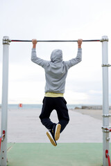 Fototapeta na wymiar Back view portrait of male runner in a sportswear pulls on the horizontal bar near the sea, strong athlete performs exercises for muscles in a cloudy day, fit man working out on street gym outdoors