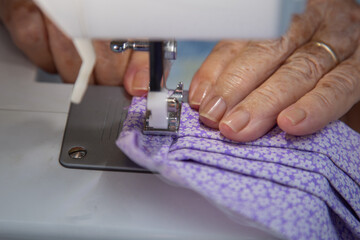 Mão de senhora idosa, usando máquina de costura para fazer máscara caseira.