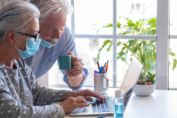 Senior man and woman with silver hair using laptop computer from home wearing face mask due to coronavirus - bright light from window
