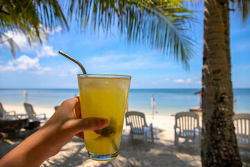 Woman hand with fresh juice on tropical beach landscape. Tropical island vacation on luxury resort