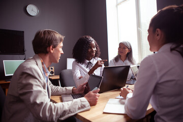 Pretty African woman speaker speaks, gesturing and pointing at someone else with his finger. Colleagues listen to her carefully. Funny business meeting in office. Toned image.