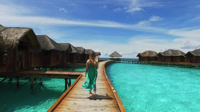 Maldive island resort. Girl in green dress walking on wooden bridge near bungalow hotel. View of a young woman with long hair from the back. Maldives travel destination. Summer vacation in paradise