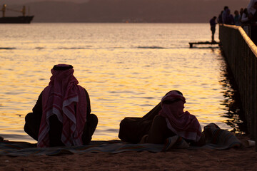 Two Arabic men wearing keffiyeh, agal and thobe are seen on the beach next to a pier. One is lying on his back the other is sitting cross legged. They look at the beautiful sunset over red sea.