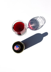 Bottle of wine and a glass with red wine casts hard shadows on a white table. The flag of the USA is depicted on the cap of the bottle.