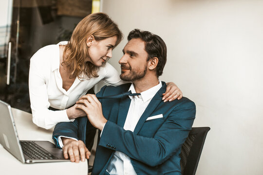 Seducing colleagues in office. Sexy blonde woman flirting with handsome smiling business man pulling him by a tie and looking into his eyes. Tinted image.
