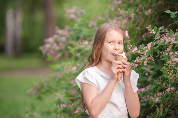 Fototapeta premium a girl with blond hair eats a delicious ice cream in a waffle cone, amid pink flowering bushes and greens, licks an ice cream