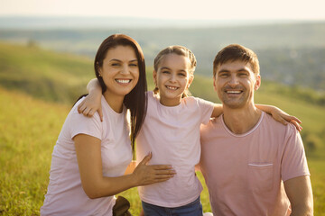 Obraz premium Portrait of positive young family looking at camera in countryside. Cute girl hugging her parents on summer vacation