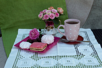 On a table on a white napkin is a bouquet of pink roses, a cup of tea, a plate on which are marshmallows, cake, candy..