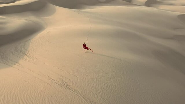 A lonely traveler in fashion red fluttering dress is walking by the beige sand dunes. 4K aerial remote view of a beautiful female model in the desert at sunset, California. Nature landscape footage