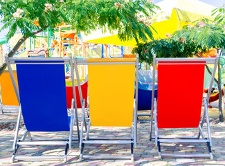 Pool loungers in 3 bright colors: blue, yellow, red standing back under green acacia trees. Mock up for summer holidays, vacation, travel concept.