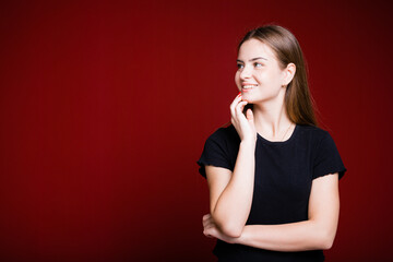Fototapeta premium Beautiful American woman in a black T-shirt holds a hand near her face and looks away smiling on a red background