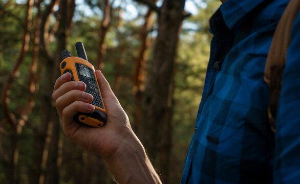 A young man in a blue plaid shirt with a walkie-talkie in his hand stands in a coniferous forest.