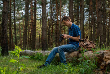 Naklejka na ściany i meble Young man in a blue plaid shirt with a camera in his hands in the coniferous forest