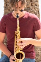 young man wearing glasses playing a saxophone while leaning on a palm tree. Street music concept.