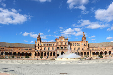 Fototapeta na wymiar Square of Spain in Seville, with the main monument palace, the fountain and a blue sky with clouds in the background - horizontal postcard or wallpaper