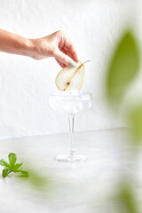 Classic martini with ice. Female hand decorates cocktail with pear slice. On a light background
