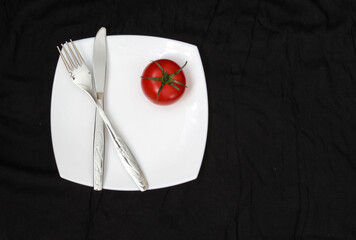 Fork, knife and white plate with tomate over black background. Clean plate and cutlery on light background. Top view.