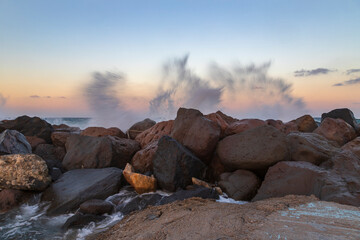 Breakwater made of stone into which waves crash. Background is blue sky at sunrise. Drops of water shatter on the stone.