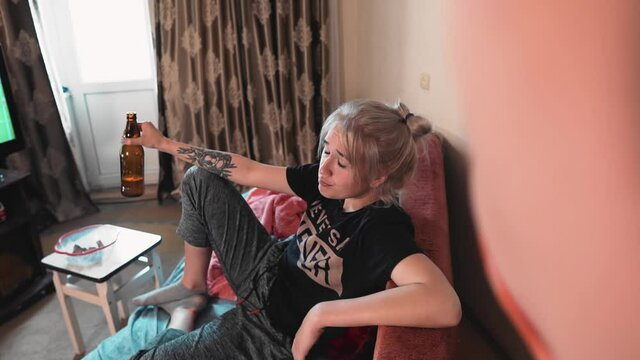A girl drinks beer sitting on the couch and a guy in an apron swears at her. The guy is henpecked.