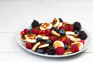Trendy mini pancakes breakfast concept. Tiny  cereal Pancakes with berries, honey or maple syrup on white background. Kids lovely breakfast concept