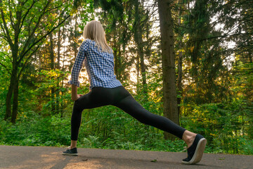 young beautiful blonde woman is engaged in exercises in the forest at sunset, making a lunge with her feet forward, rear view