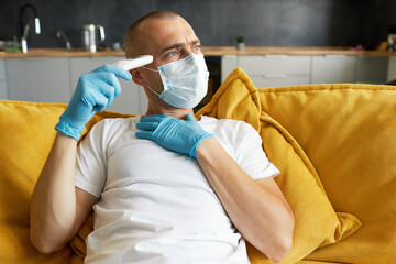 A sick man in a protective mask lies at home on the sofa and holds his hand on his forehead during quarantine. Measures temperature with a hand and waits for a doctor