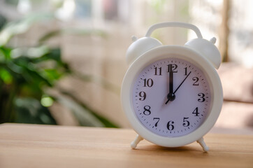 White alarm clock on a wooden table in the sun in a room with indoor plants, copy space