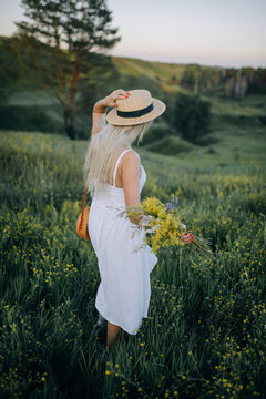 Girl in a white summer dress and hat with flowers in her hands on a background of beautiful nature on the hill.Vertical photo