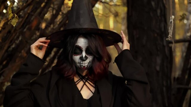 Portrait of a witch in a hat cap smiling and flirting in nature. Theme of costumes and transformation for holiday of Halloween. Traditional skull makeup for day of the dead Santa Muerte celebration