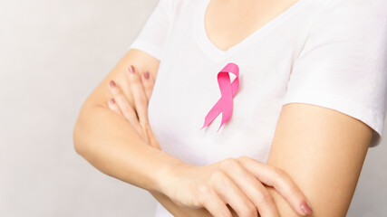 Portrait of woman standing crossed arms over chest, wear white t-shirt with pink ribbon pin as a symbol of support on breast cancer day campaign.