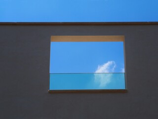 View of single cloud in blue sky through a window frame on gray wall of modern building