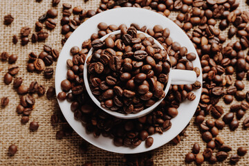 White cup fulled of roasted coffee beans on sackcloth background, close up