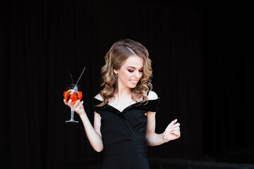 Beautiful blonde, young girl holding an aperol spritz. Aperol cocktail syringe in a glass. Portrait of a young girl in a restaurant. Young girl in a restaurant with a glass on a black background.