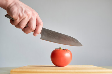 Hand with a knife, about to cut a ripe tomato on a wooden chopping board.