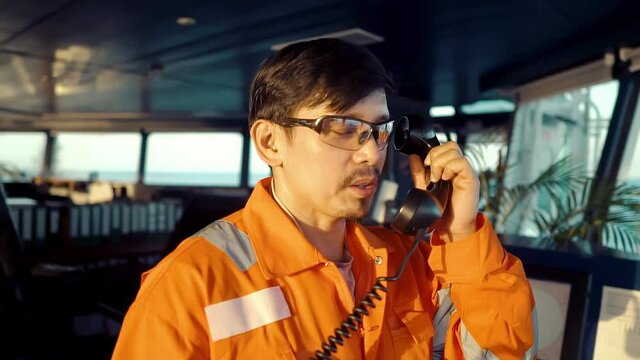 Filipino deck Officer on bridge of vessel or ship wearing coverall during navigaton watch at sea . He is speaking on GMDSS VHF radio, communication between vessels.