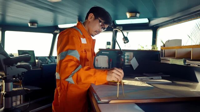 Filipino deck Officer on bridge of vessel or ship wearing coverall during navigaton watch at sea . He is plotting position on chart
