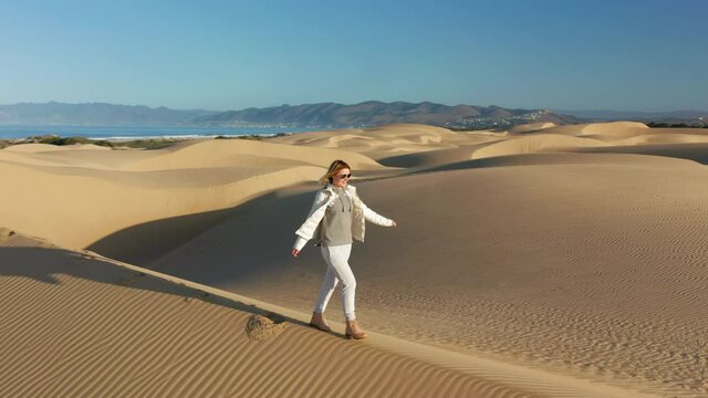 Happy young woman in white jacket is cheerfully raising hands up. Joyful model in the desert nature. 4K circular aerial overhead video of a traveler running by the golden sand dunes, California, USA