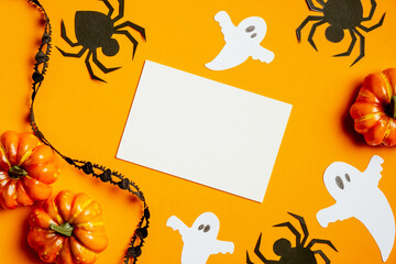 Halloween greeting card mockup with pumpkins, black spiders, ghosts, ribbon on orange background. Flat lay, top view. Happy Halloween concept.