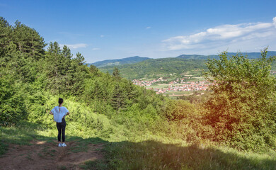 Naklejka na ściany i meble Back view of young woman standing on top of a hill. Attractive girl enjoying the colorful countryside view. Small village at the foot of a hill, beautiful landscape with blue sky.