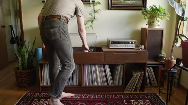 Barefooted hipster man listening to music walks up to a vintage stereo to flip a vinyl record